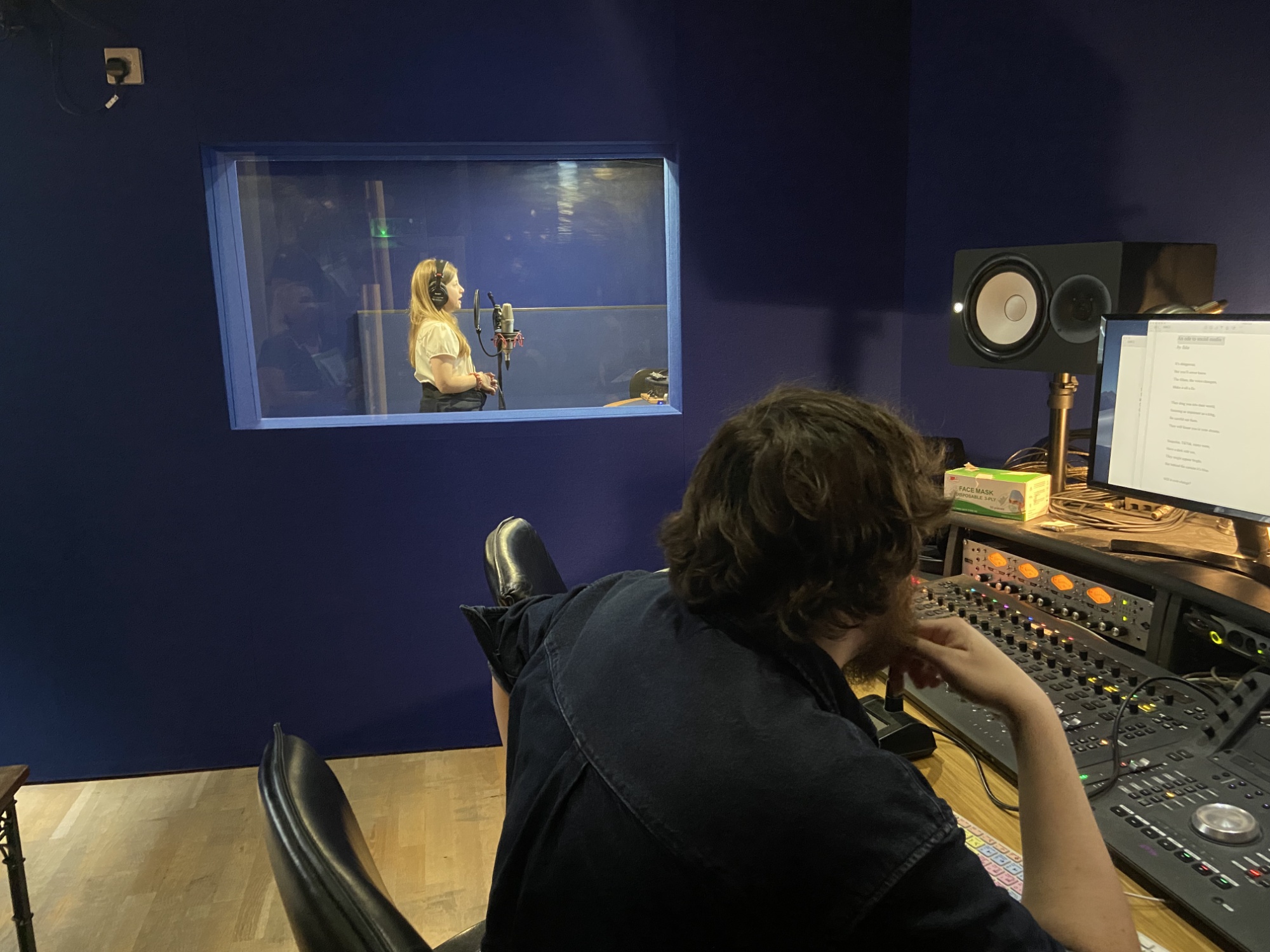 In the recording studio - a child in school uniform stands in a soundproof booth wearing headphones and speaking into a microphone.